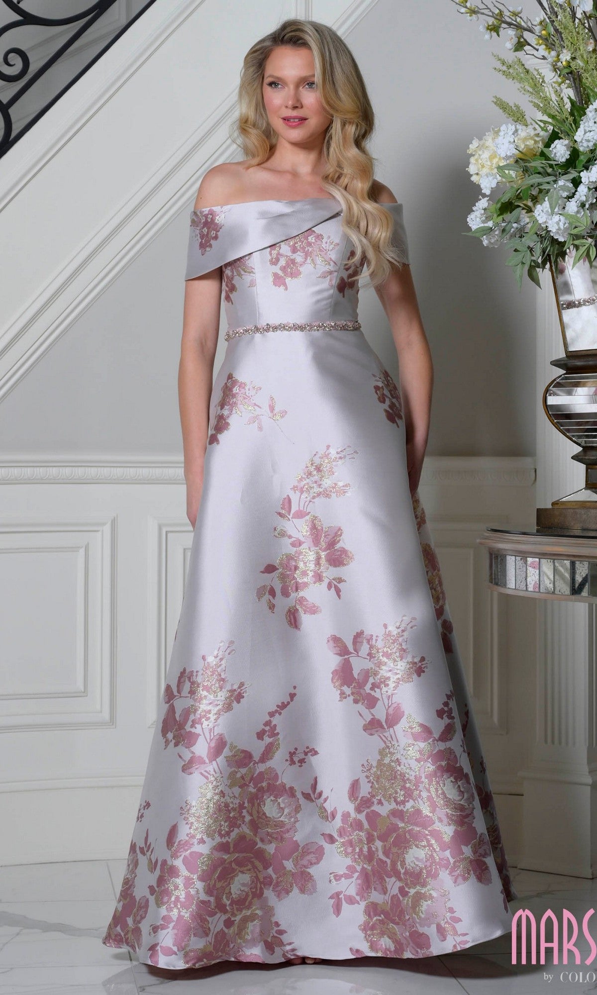 A woman stands indoors near a staircase and floral arrangement, wearing the Marsoni MV1402 Long Formal Dress in Rose Multi, featuring jacquard fabric with pink floral patterns and an off-the-shoulder design.